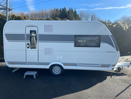 Weißer
                           Wohnwagen mit grauem Seitenstreifen in Seitenansicht auf einem asphaltierten Platz. Zu sehen sind Eingangstür, Fenster, Lüftungsgitter,
                           Rad, Deichsel und eine kleine Einstiegshilfe vor der Tür.