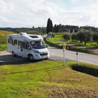 Ein weißes Wohnmobil parkt auf einem unbefestigten Seitenstreifen direkt an einer asphaltierten Landstraße. Die Beifahrertür
                           steht weit offen. Im grasgrünen Vordergrund stehen Begrenzungspfähle, einer davon trägt ein gelbes Schild mit roten Pfeilen.
                           Im Hintergrund erstreckt sich eine weite toskanische Hügellandschaft mit herbstlich gefärbten Weinbergen, Olivenbäumen und
                           hoch aufragenden, dunklen Zypressen. Ein blaues Straßenschild am rechten Fahrbahnrand weist auf den Ort „Castelnuovo Berardenga“
                           hin. Darüber spannt sich ein blauer Himmel mit hellen Quellwolken.