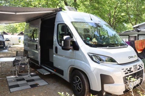 Wohnmobil Weinsberg CaraLife am Campingplatz mit Markise, Campingtisch,
                           Sessel und Teppich zwischen grünen Baumen.