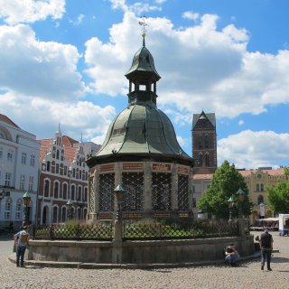 Ein detaillierter
                           Blick auf die Wismarer Wasserkunst, ein kunstvoller, pavillonartiger Brunnenbau aus der Renaissance im Zentrum des kopfsteingepflasterten
                           Marktplatzes. Das Bauwerk hat ein grünes Kupferdach mit einer kleinen Laterne und ist von einem schmiedeeisernen Zaun umgeben.
                           Im Hintergrund sind historische Giebelhäuser und der Turm der Marienkirche unter einem bewölkten Sommerhimmel zu sehen.
