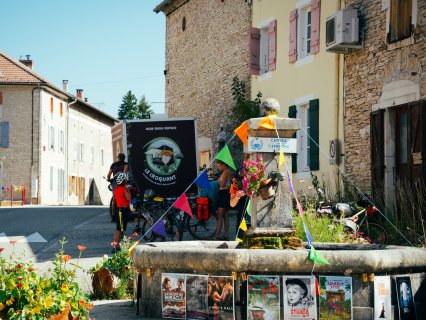 Ein steinerner Brunnen
                           mit Blumen und bunten Wimpeln steht an einer Dorfstraße. Daneben befinden sich mehrere bepackte Fahrräder und Personen. Im
                           Hintergrund sind Steinhäuser, ein Food-Truck-Schild und mehrere Plakate am Brunnenrand zu sehen.