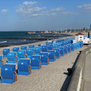 Ein breiter Sandstrand im Ostseebad
                           Kühlungsborn, auf dem zahlreiche leuchtend blaue Strandkörbe in sauberen Reihen aufgestellt sind. Rechts verläuft die gemauerte
                           Strandpromenade mit einer weißen Verkaufs- und Rettungsstation. Im Hintergrund ist die Hafeneinfahrt des Bootshafens mit vielen
                           Segelbootmasten unter leicht bewölktem Himmel zu sehen.