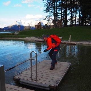 Person mit Rucksack steht auf einem kleinen Holzfloß in einem Teich
                           vor verschneiten Bergen.