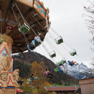 Kettenkarussell mit grünen Sitzen vor herbstlichen
                           Bäumen und verschneiten Bergen.