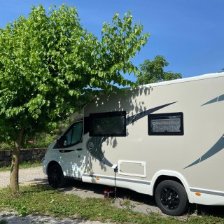 Ein modernes, weißes Wohnmobil mit der Modellnummer „640“ steht auf einem geschotterten Camping-Stellplatz, der
                           von einer grasbewachsenen Böschung und einem jungen Laubbaum umrahmt wird. Im Vordergrund steht ein kleiner, ausgeklappter
                           Holztisch mit einer Flasche Wein und zwei Gläsern. Direkt daneben befindet sich ein Holzschild mit der Aufschrift „PARZELLE
                           14“ und der Unterschrift „Sakrida Vineyard Camping - Ecocampming Resort“. Im Hintergrund erstrecken sich weitläufige, akkurat
                           angelegte Weinberge auf sanften Hügeln unter einem strahlend blauen, fast wolkenlosen Himmel.