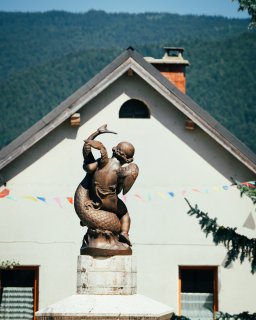 Eine bronzefarbene Statue einer geflügelten Kinderfigur
                           mit einem großen Fisch steht auf einem hellen Sockel vor einem Gebäude mit Giebeldach. Im Hintergrund sind bewaldete Berge
                           zu sehen, vor dem Gebäude hängen bunte Wimpelgirlanden.