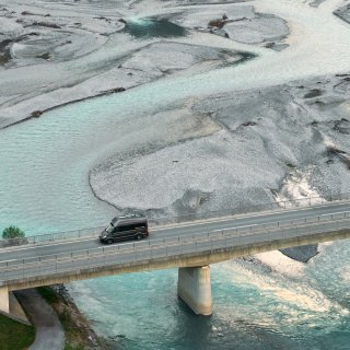 Ein Camper überquert eine Brücke über einen Fluss mit türkisfarbenem Wasser und weitläufigen Kiesbänken.
                           Die Szene ist aus der Vogelperspektive aufgenommen.