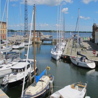 Blick
                           von oben auf einen Kanal im Hafen von Stralsund, in dem zahlreiche Segelboote und Motoryachten dicht an dicht an den Stegen
                           vertäut sind. Rechts verläuft eine gepflasterte Kaimauer mit Sitzgelegenheiten im Freien, während sich im Hintergrund eine
                           Brücke und das offene Wasser des Strelasunds unter blauem Himmel erstrecken.