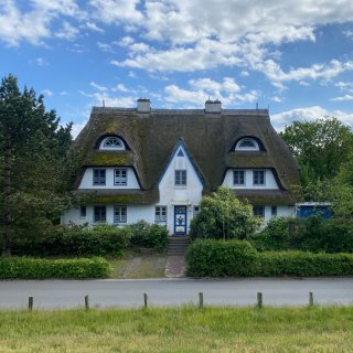 Ein traditionelles,
                           großes Reetdachhaus im Ostseeheilbad Zingst. Das weiße Gebäude besitzt blau abgesetzte Fensterrahmen, eine kunstvoll verzierte
                           Eingangstür und charakteristische Fledermausgauben im dicken, moosbesetzten Schilfrohrdach. Vor dem Haus verläuft eine asphaltierte
                           Straße, die durch eine niedrige Hecke und Holzpfähle von einer Rasenfläche getrennt ist.