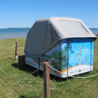 Ein spezieller, geschlossener
                           Schlafstrandkorb steht auf einer grünen Wiese direkt an der Küste von Zierow. Der Korb ist mit einer grauen Wetterplane abgedeckt
                           und an den Seiten mit einem Dünenmotiv bedruckt. Ein Seilzaun grenzt den Bereich ein, im Hintergrund erstreckt sich das ruhige,
                           blaue Meer unter wolkenlosem Himmel.