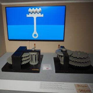 Auf einem hellgrauen Präsentationstisch stehen zwei mechanische Apparaturen mit massiven schwarzen Grundplatten und Rahmen.
                           In diese sind graue, gestufte Zahnräder und zylinder- sowie kegelförmige Konstruktionen eingespannt, die jeweils über eine
                           vertikale Handkurbel mit kleinem Holzgriff angetrieben werden. Zwischen den beiden Objekten liegt ein kleines Hinweisschild
                           mit einem durchgestrichenen Hand-Symbol. Im unmittelbaren Vordergrund verläuft eine flache Infotafel mit Erklärtexten und
                           historischen Skizzen. An der beigefarbenen Wand im Hintergrund hängt ein breiter Flachbildschirm, der das digitale 3D-Modell
                           eines weißen Schneckengetriebes vor einer leuchtend blauen Fläche zeigt.