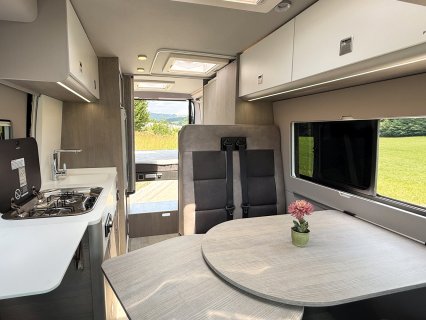 Blick in den hellen Innenraum des Furgok Xpace 600 in Richtung Heck. Links sieht man die Küchenzeile
                           mit hochgeklappter Herdabdeckung, rechts einen Tisch und eine Sitzbank mit Anschnallgurten. Im Hintergrund blickt man durch
                           die offenen Hecktüren ins Grüne.