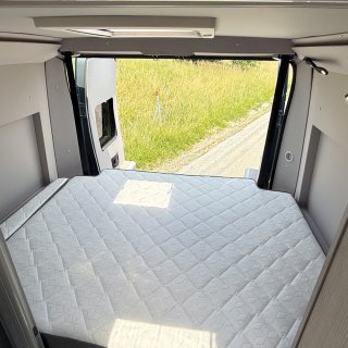 Man blickt aus dem
                           Inneren des Campers direkt über die breite, weiß gesteppte Matratze des Heckbetts. Die Hecktüren stehen weit offen und geben
                           den Blick auf einen Feldweg und eine sonnige, grüne Wiese frei. Oben ist noch ein Dachfenster zu erkennen.