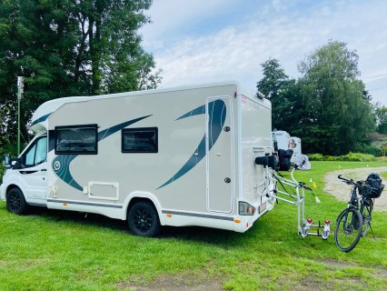 Ein weißes Wohnmobil
                           steht auf einer grünen Rasenfläche. Am Heck befindet sich ein ausgeklappter Fahrradträger, daneben steht ein schwarzes Fahrrad
                           mit Gepäcktasche. Im Hintergrund sind weitere Wohnmobile, Bäume und ein geschotterter Fahrweg zu sehen.