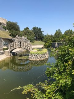 Das Bild zeigt einen Teil des Ferrari-Gartens in Štanjel, Slowenien. Man
                           sieht einen Fluss, über dem eine kleine steinerne Brücke drüber geht. Im Hintergrund sieht man Wiesen, Bäume und weitere steinerne
                           Bauten.