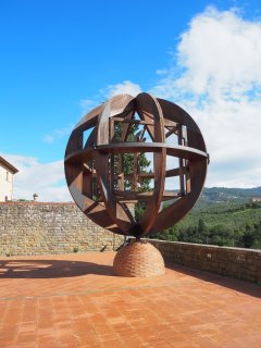 Eine große, kugelförmige Skulptur aus massiven, gebogenen
                           Holzbalken, die an den Schnittstellen mit metallischen Beschlägen fixiert sind, steht im Zentrum der Aufnahme. Im Inneren
                           der offenen Kugel bilden gerade Streben ein komplexes geometrisches Gerüst. Das Kunstwerk ruht auf einem kegelförmigen Sockel
                           aus geschichteten, rötlichen Ziegelsteinen. Der weite Platz ist mit quadratischen Terrakotta-Fliesen gepflastert, auf die
                           die Konstruktion markante Schatten wirft. Im Hintergrund rahmen eine niedrige Natursteinmauer, ein angeschnittenes Gebäude
                           auf der linken Seite und dichtes Grün den Blick auf eine bewaldete Berglandschaft unter einem leuchtend blauen, leicht bewölkten
                           Himmel ein.