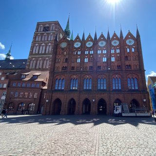 Die imposante Schaufassade des
                           historischen Stralsunder Rathauses aus rotem Backstein am Alten Markt unter strahlendem Sonnenschein. Das gotische Gebäude
                           beeindruckt mit einer reich verzierten Schmuckwand, spitz zulaufenden Türmchen und kreisrunden Fensteröffnungen. Links im
                           Hintergrund ragt der mächtige Turm der St.-Nikolai-Kirche empor.
