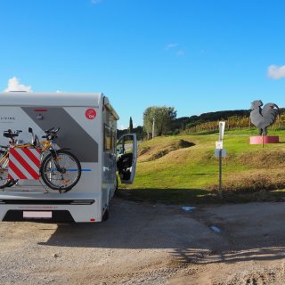 Ein weißes Wohnmobil parkt auf
                           einem unbefestigten, teils von Pfützen durchzogenen Schotterplatz am Straßenrand. Die Seitentür der Kabine steht offen. Am
                           Heck des Fahrzeugs ist ein Fahrradträger mit montierten Rädern und einer markanten, rot-weiß gestreiften Warntafel angebracht.
                           Auf einem leichten, grünen Grashügel rechts daneben steht eine große, dunkle Metallskulptur in Form eines Hahns, die auf einem
                           rosafarbenen, runden Sockel fixiert ist. Im Hintergrund erstrecken sich weitläufige Weinberge und einige Bäume unter einem
                           strahlend blauen, fast wolkenlosen Himmel.