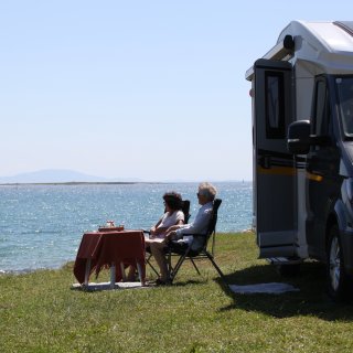 Zwei Personen sitzen auf Campingstühlen neben einem Wohnmobil am Ufer eines Gewässers. Vor ihnen steht
                           ein kleiner Tisch, während das Fahrzeug auf einer Wiese direkt am Wasser parkt.
