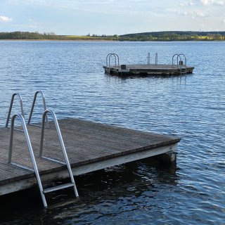 Ein hölzerner Badesteg
                           mit Metallleiter im klaren Wasser eines Sees beim Camping Resort Havelberge. Im Hintergrund schwimmt eine Badeinsel auf dem
                           ruhig fließenden Wasser, dahinter das grüne, bewaldete Seeufer unter leicht bewölktem Himmel.