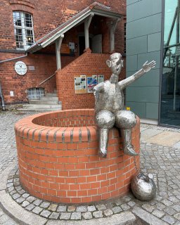 Metallfigur mit ausgestrecktem Arm auf dem
                           Rand eines runden gemauerten Brunnens. Neben der Figur liegt eine kugelförmige Metallskulptur an einer Kette. Im Hintergrund
                           befindet sich der Eingang eines roten Backsteingebäudes mit einem Schild des Spreewald-Museums.