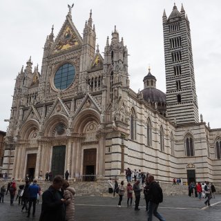 Ein Foto der prächtigen
                           Kathedrale von Siena aus einer seitlichen Perspektive, die die markanten schwarz-weiß gestreiften Marmormauern, den Campanile
                           und die Kuppel hervorhebt. Im Hintergrund ist die aufwendige Westfassade zu sehen. Auf der Piazza davor, seitlich geparkt,
                           steht ein weißes Wohnmobil, aus dessen Fenster eine Person schaut. Der Platz ist voller Touristen unter einem bewölkten Himmel.