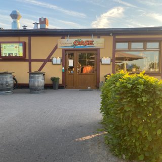 Die Außenansicht
                           des Restaurants „Oase mit Kegelbahn“ im Ostsee-Campingpark Zierow. Das gelbe Gebäude in Fachwerk-Optik hat eine hölzerne Eingangstür
                           und große Fenster. Auf dem gepflasterten Vorplatz stehen zwei große Holzfässer als Stehtische, im Vordergrund ist ein grüner
                           Busch zu sehen.