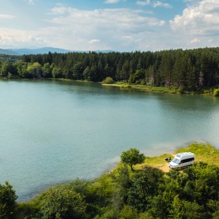 Ein Camper steht auf einer kleinen Lichtung direkt am Ufer eines Sees. Die Luftaufnahme zeigt die umgebenden
                           Bäume, den Wald am gegenüberliegenden Ufer und das weitläufige Gewässer.