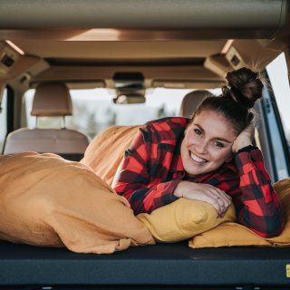 Eine Person liegt auf einem Bett im Innenraum eines Campers und stützt den Kopf auf eine Hand. Mehrere
                           Kissen und eine Decke befinden sich auf der Liegefläche