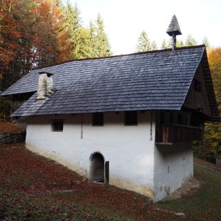 Historisches Bauernhaus mit weiß verputztem
                           Untergeschoss, hölzernem Aufbau und Schindeldach im herbstlichen Wald.