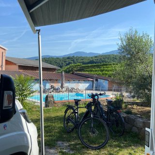 Der Blick fällt aus dem Schatten einer ausgefahrenen grauen Wohnmobil-Markise auf die angrenzende Parzelle. Links
                           im Vordergrund ist die weiße Front des Wohnmobils angeschnitten. Auf der Wiese davor parken zwei schwarze E-Bikes. Direkt
                           dahinter befindet sich ein umzäunter, hellblauer Swimmingpool mit Sonnenliegen. Im Hintergrund sind ein Gebäude mit bunt bemalter
                           Wand, saftig grüne Weinberge und eine weite Bergkulisse bei strahlend blauem Himmel zu sehen.