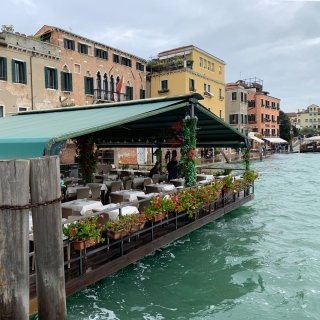 Überdachte Restaurant-Terrasse direkt über dem Wasser eines venezianischen Kanals. Das Plateau wird
                           von einem grünen Zeltdach geschützt und ist an den Rändern mit roten Blumen in Pflanzkästen sowie rankenden Blättern an den
                           Stützpfeilern dekoriert. Darunter stehen eingedeckte Tische mit weißen Tischdecken. Im linken Vordergrund ragen dicke, verwitterte
                           Holzpfähle mit einer eisernen Kette aus dem türkisgrünen Wasser. Im Hintergrund säumen bunte, historische Häuserfassaden in
                           Ziegelrot, Gelb und Pfirsichfarben die gepflasterte Uferpromenade unter einem bewölkten Himmel.
