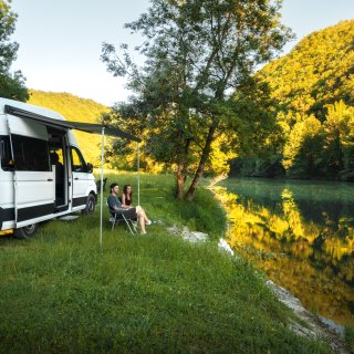 Zwei Personen sitzen auf Campingstühlen neben einem Camper mit ausgefahrener Markise. Das Fahrzeug steht
                           auf einer Wiese am Ufer eines Gewässers, umgeben von Bäumen und bewaldeten Hängen.