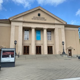 Die klassizistische
                           Fassade des Landestheaters Neustrelitz mit großen Säulen und einer Inschrift im Giebel unter blauem Himmel. Vor den Eingangsstufen
                           befindet sich ein gepflasterter Vorplatz mit einem Schaukasten für aktuelle Schauspiel-Aufführungen.