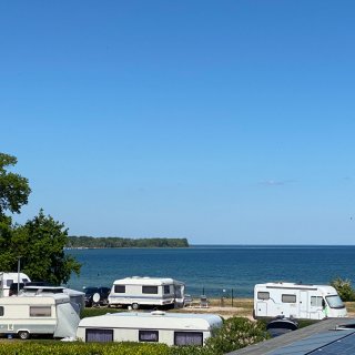 Blick über mehrere abgestellte Wohnwagen und Wohnmobile
                           im Ostsee-Campingpark Zierow direkt auf das blaue Meer der Wismarer Bucht. Am Horizont ist eine bewaldete Landzunge unter
                           einem strahlend blauen, wolkenlosen Himmel zu sehen.