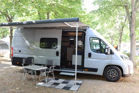 Wohnmobil Weinsberg CaraLife am Campingplatz mit Markise, Campingtisch,
                           Sessel und Teppich zwischen grünen Baumen.
