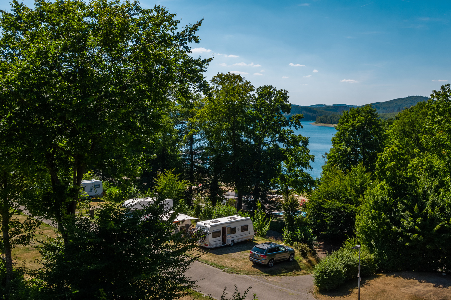 Foto von mit Autos und Wohnwägen besetzten, von Bäumen umgebenen Park-/Stellplätzen vor dem Biggesee.