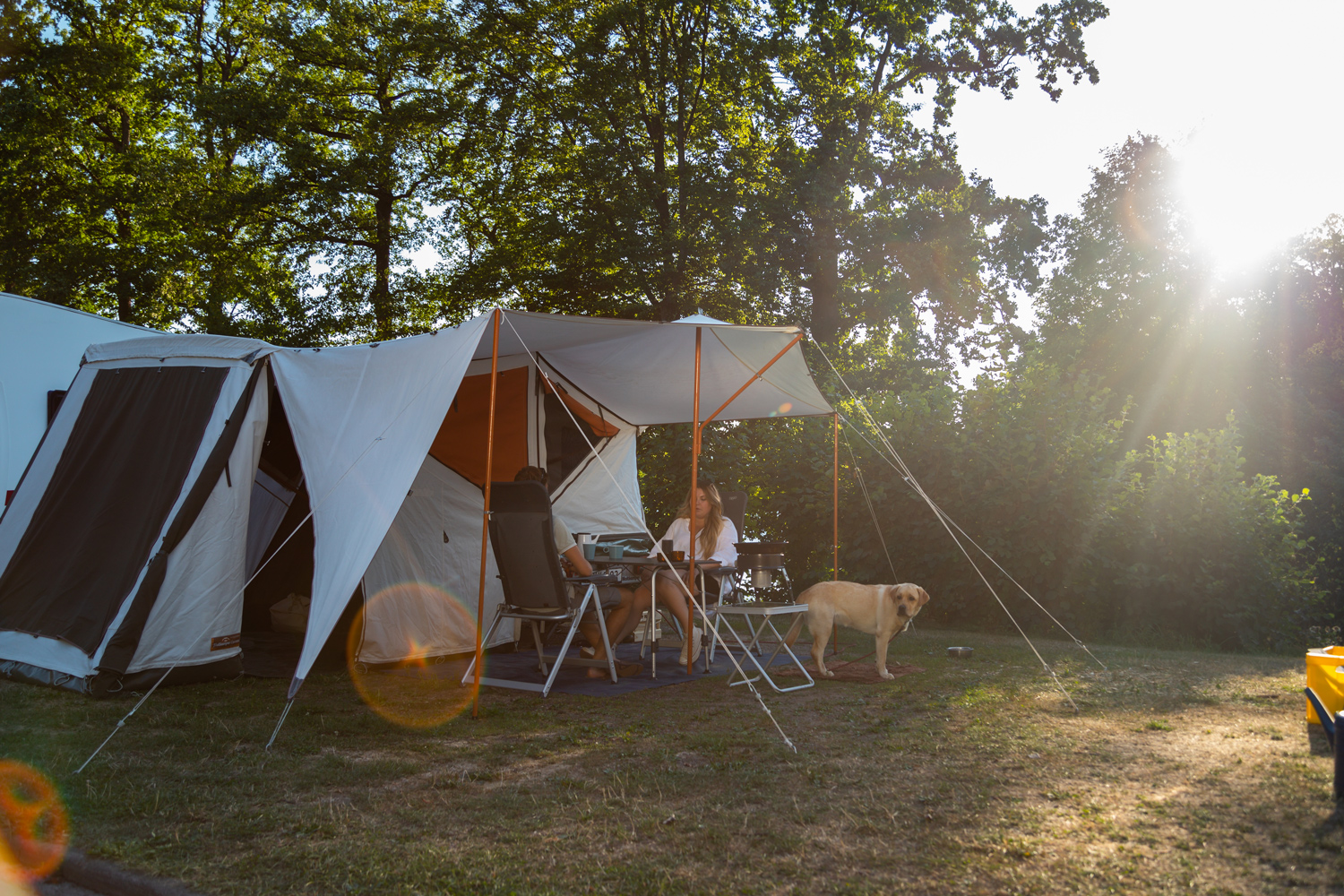 Zwei Personen
                                 sitzen an Campingtisch vor Zelt auf Wiese mit Hund daneben.