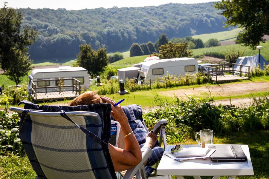 Person,
                                 welche auf Campingstuhl und neben Campingtisch sitzt und eine Zeitschrift liest. Im Hintergrund sieht man Wohnwägen, sowie
                                 Wiesen und Wald. 