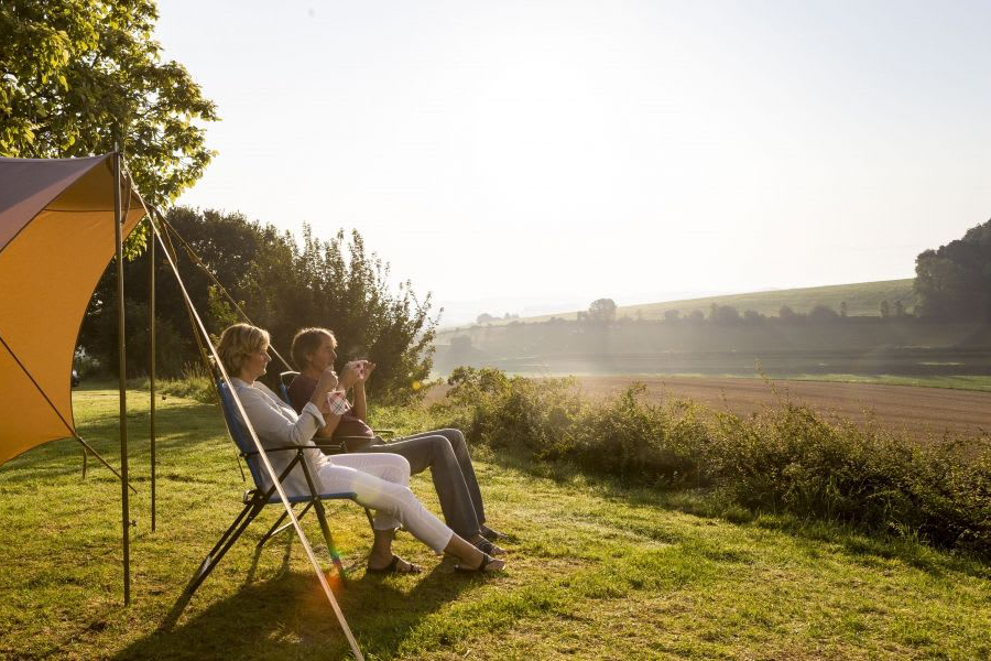 Zwei Personen, welche auf Campingstühlen vor einem Zelt sitzen mit Tassen in der Hand und in
                                 ein Feld schauen. 