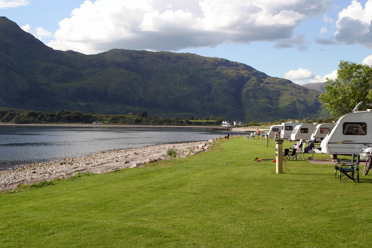 Auf dem Bild sind Stellplätze vom Campingplatz Bunree in Großbritannien
                                 abfotografiert. Man sieht Wohnwägen und Mobile, welche auf einer Wasser vor einem kleinen See parken und jeweils Campingstühle
                                 vor den Fahrzeugen.