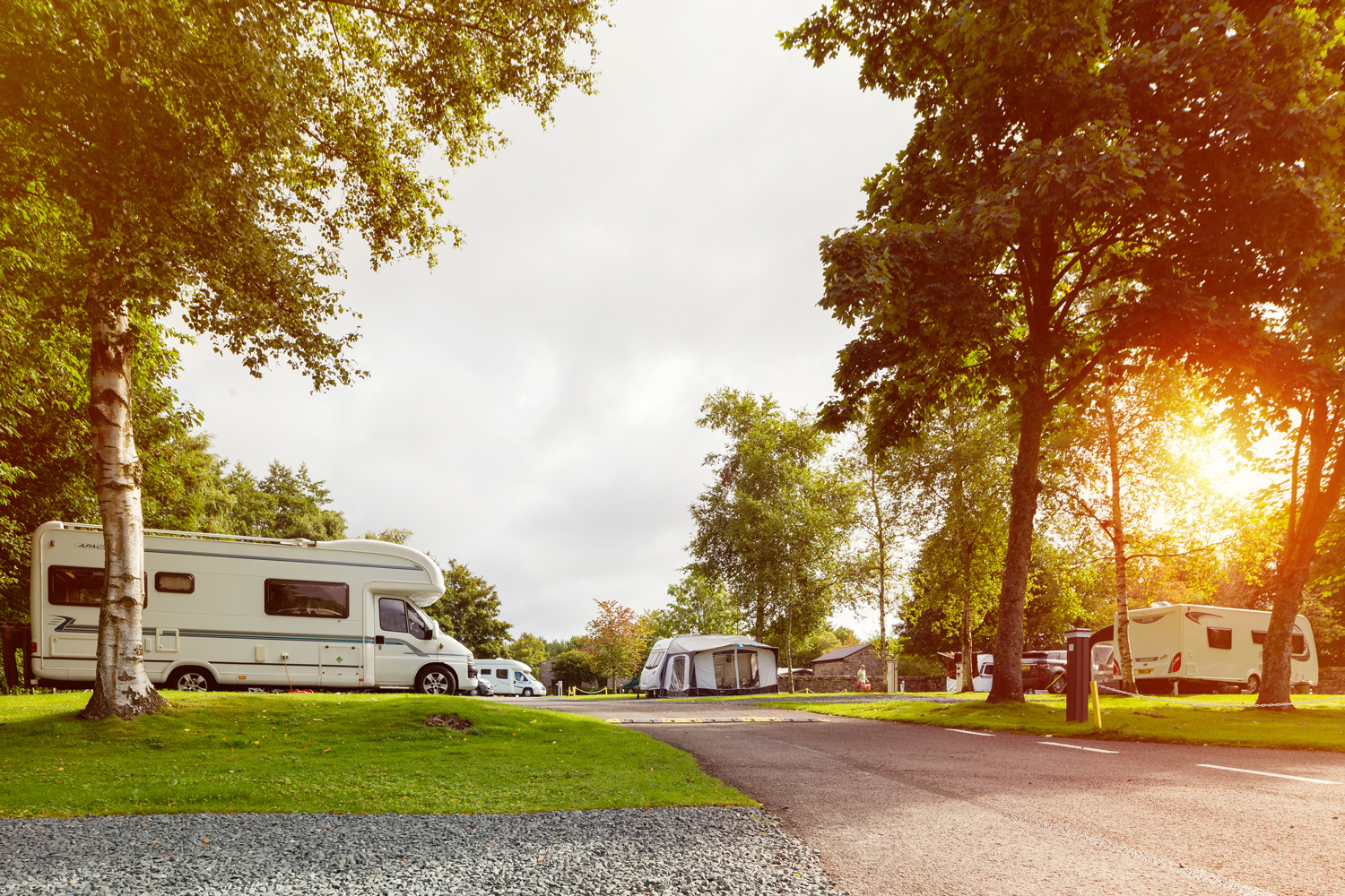 Auf dem Bild sind Stellplätze vom Campingplatz Hawes in Großbritannien abfotografiert. Man sieht Wohnmobile,
                                 sowie einen Wohnwagen mit Zelt, welche auf einer Wiese unter Bäumen stehen.