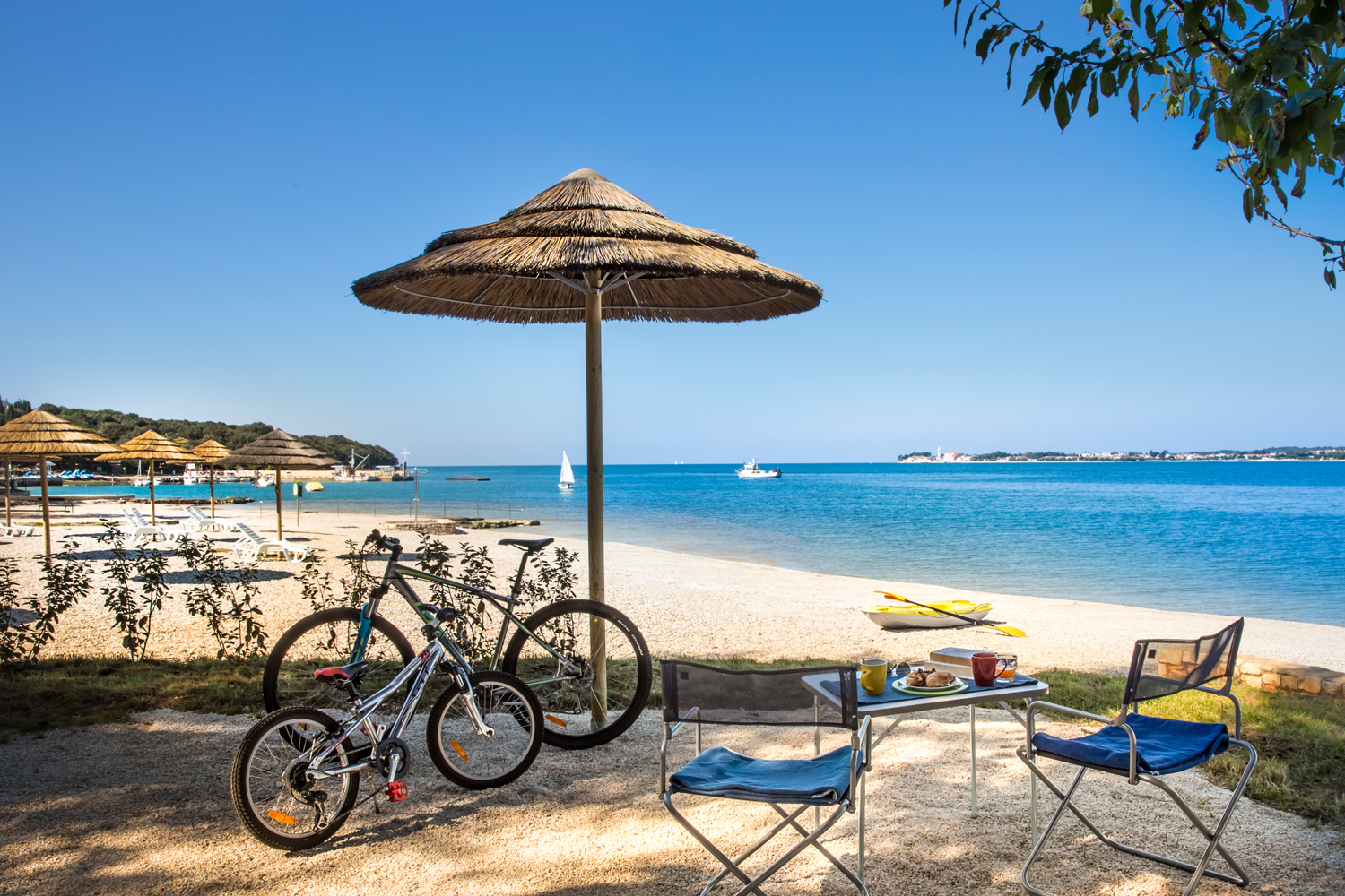Auf dem Bild sieht man zwei Campingstühle und einem Campingtisch auf einem Strand, im Schatten vor einem Holzschirm
                                 und neben zwei Fahrrädern. Im Hintergrund sieht man den Rest vom Strand mit einem Boot und das Meer mit Segel- und Fischerboot,
