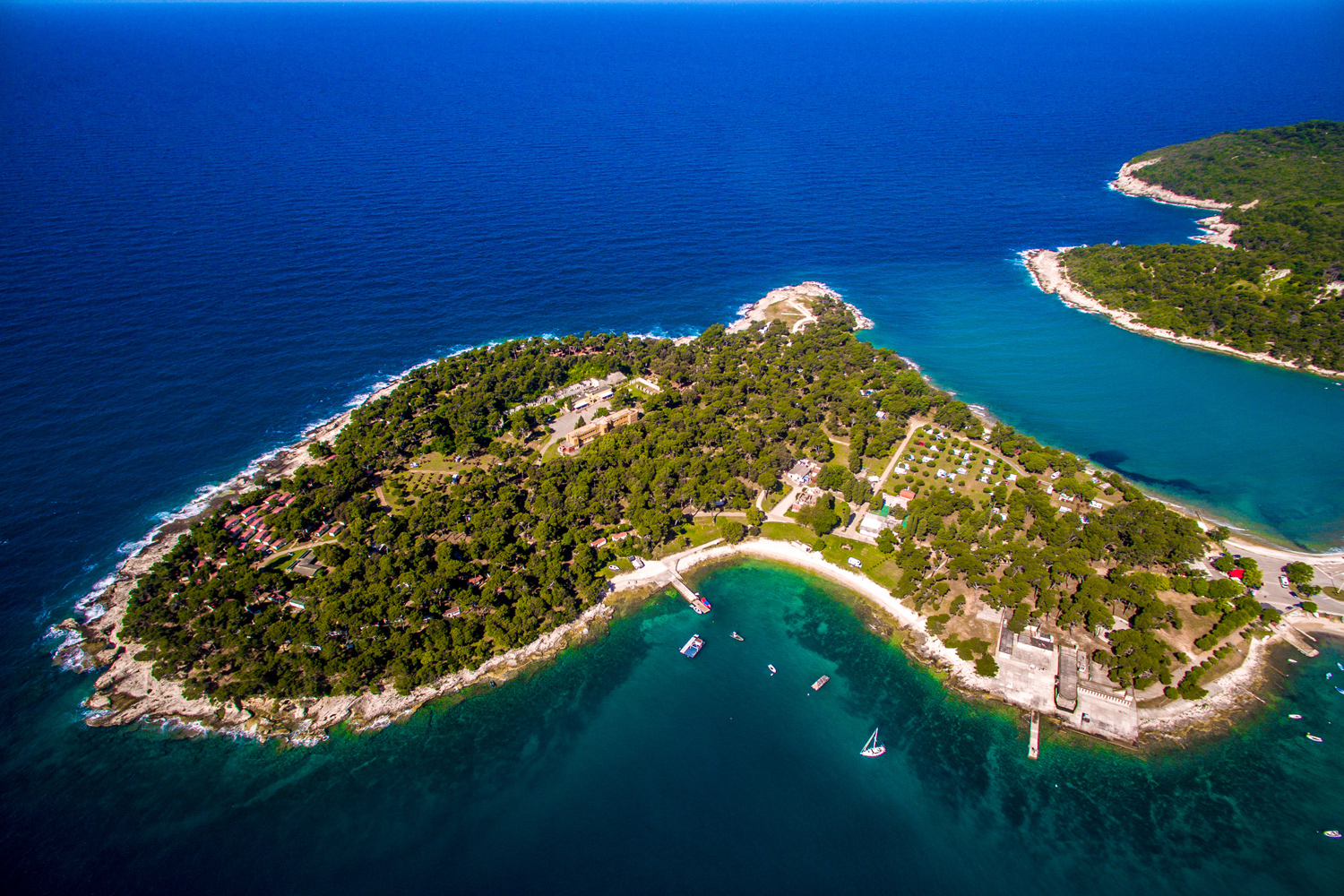 Luftbildaufnahme der
                                 Arena Stoja Campsite. Ein Campingplatz auf einer Halbinsel, welche mit vielen Bäumen bewachsen und einem kleinen Hafen mit
                                 Booten davor.