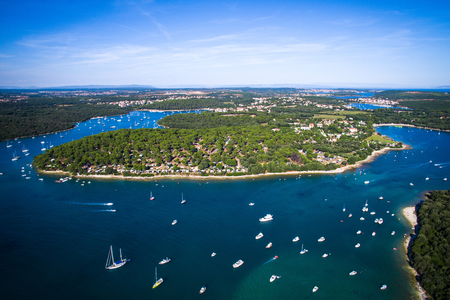 Luftbildaufnahme des Arena Indije Campsite. Der Campingplatz liegt direkt am Wasser und ist stark mit Bäumen
                                 bewachsen. Das Wasser vor dem Platz ist voll mit kleinen und großen Booten, von Segelbooten bis hin zu Yachten.