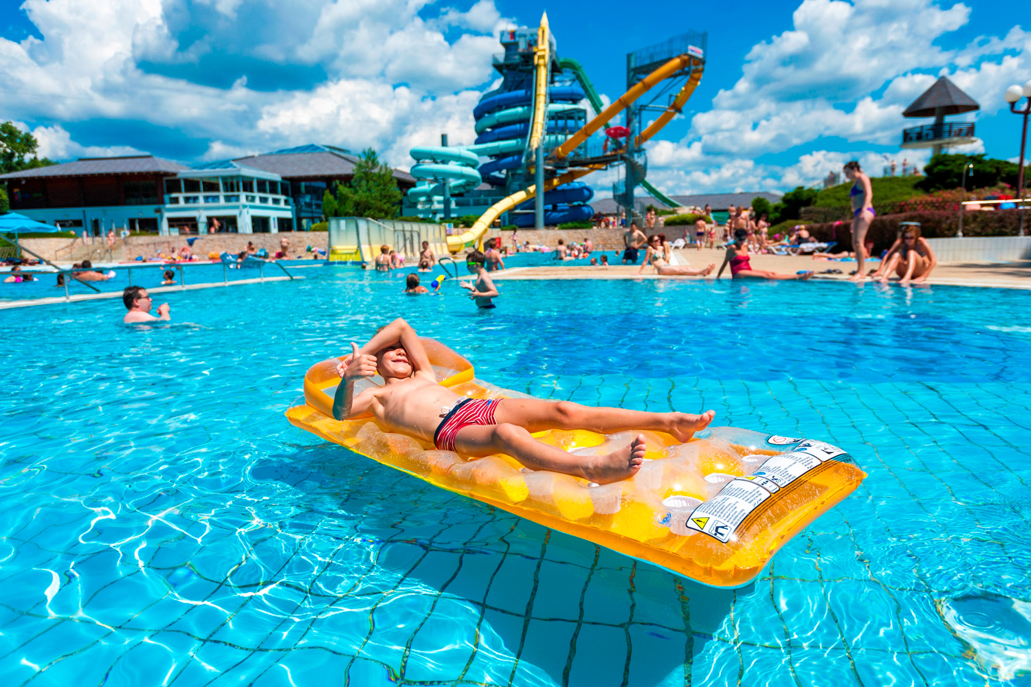 Auf dem Bild sieht man ein Kind, welches auf einer Luftmatratze in einem
                                 Schwimmbecken des Sava Moravske Toplice Camping Terme 3000 liegt. Im Hintergrund sieht man die Rutschen der Anlage sowie einen
                                 vollen Pool. 