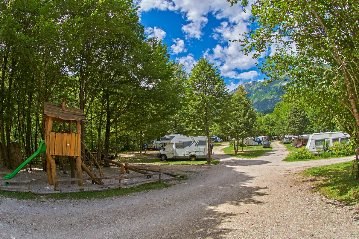 Foto des Eco Campingplatzes Kamp Koren in Slowenien. Die Straße auf dem
                                 Platz ist ein Schotterweg und die Plätze sind auf Gras. Man sieht einige Wohnmobile und Wohnwägen am Platz stehen. Der Platz
                                 ist stark mit Bäumen bewachsen und auf der rechten Seite des Bildes ist ein Kinderspielplatz mit Rutsche. 