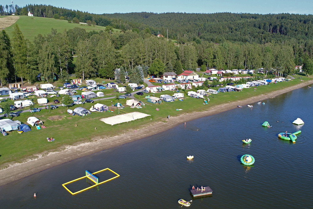 Luftbildaufnahme des Camping Resort Frymburk in Tschechien. Der Campingplatz liegt auf einer Wiese direkt
                                 am Wasser und hat u.A. einen Beachvolleyball-Platz sowie mehrere Spiele im Wasser. Der Platz ist von dichtem Wald umgeben.
                                 