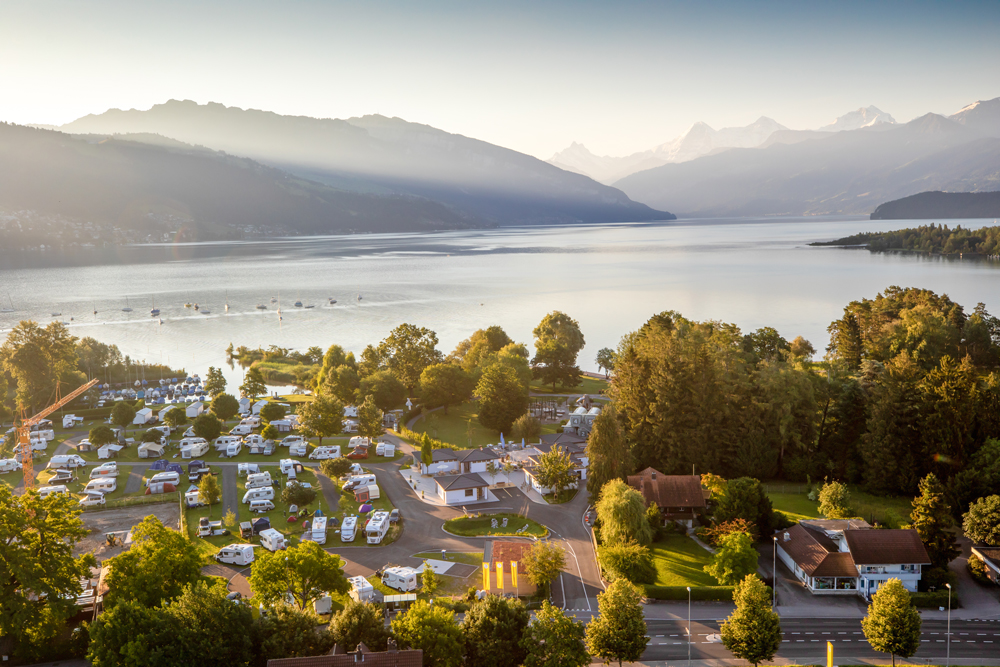 Drohnenaufnahme bei Sonnenuntergang des TCS Camping Gwatt
Thunersee in Schweiz. Auf dem Bild sind die belegten Plätze des Platzes zu sehen, welche direkt am See liegen mit Bergpanorama
im Hintergrund.
