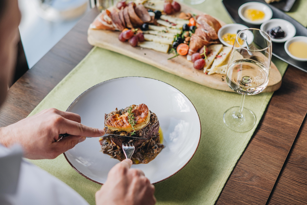 Person, welche am Tisch im Steakhouse
des Resorts San Servolo sitzt von hinten abfotografiert. Die Person schneidet gerade ein Stück Fleisch auf und vor ihm auf
dem Tisch liegt zudem eine Platte mit Käse, Schinken, usw.